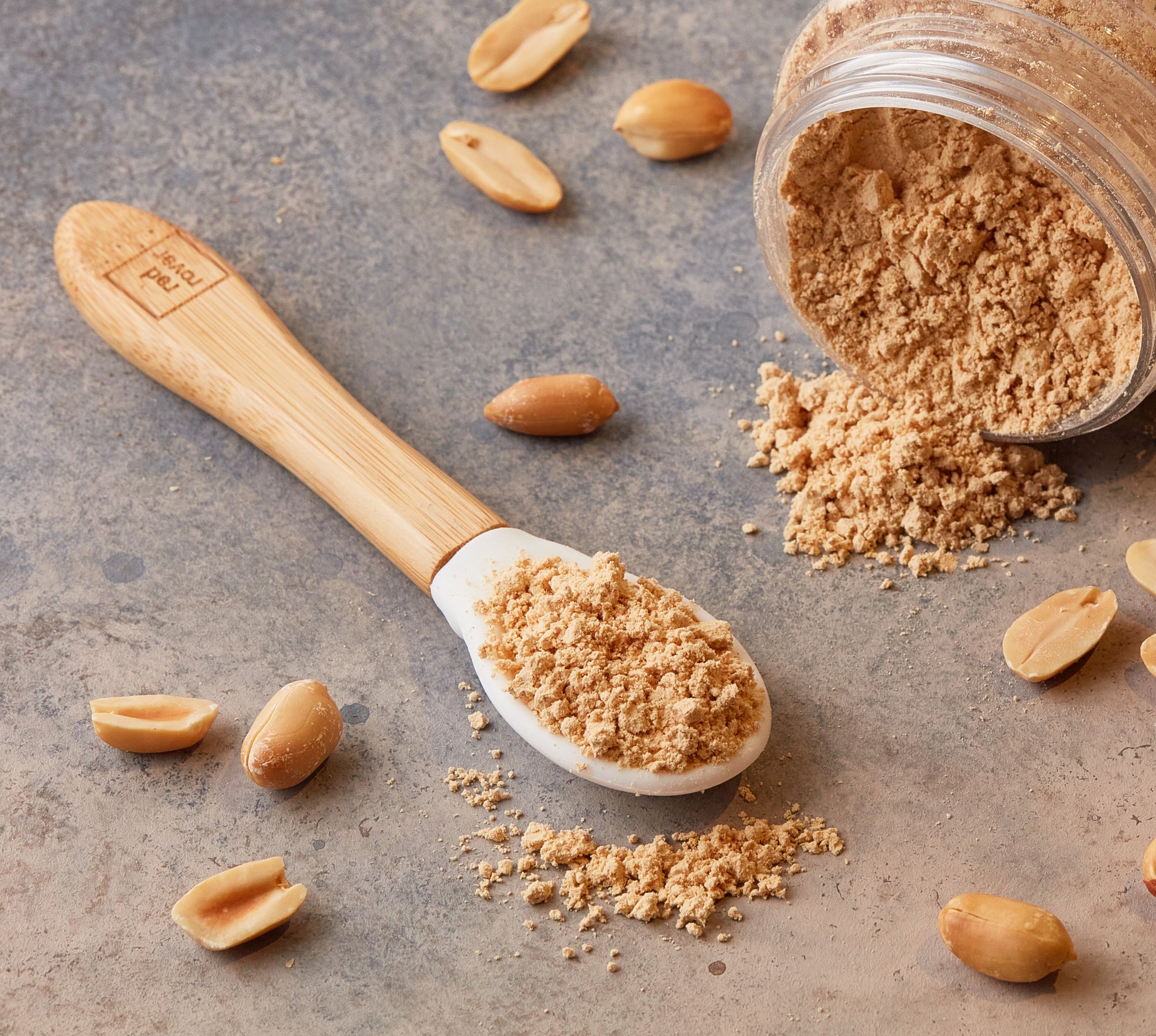 pb2 jar on side spilling onto table with spoon with powdered peanut butter on it. Peanut pieces scattered on table. 