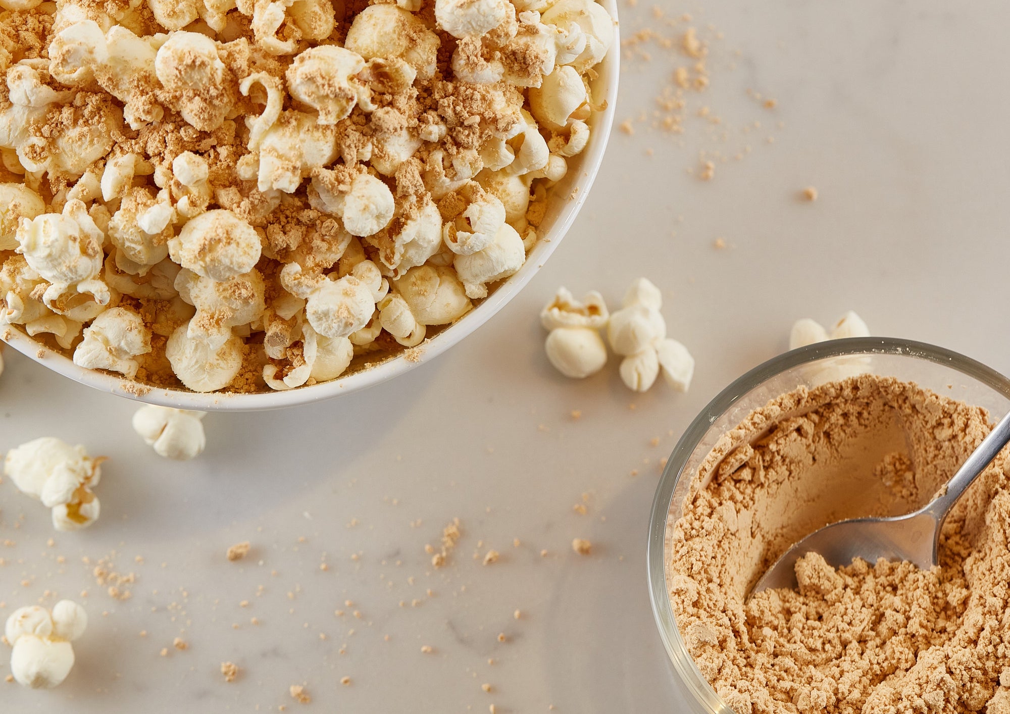 Popcorn in a bowl with PB2 powdered peanut butter sprinkled on it. Along with a small bowl of powdered peanut butter with a spoon in it. 