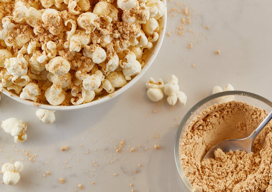 Popcorn in a bowl with PB2 powdered peanut butter sprinkled on it. Along with a small bowl of powdered peanut butter with a spoon in it. 